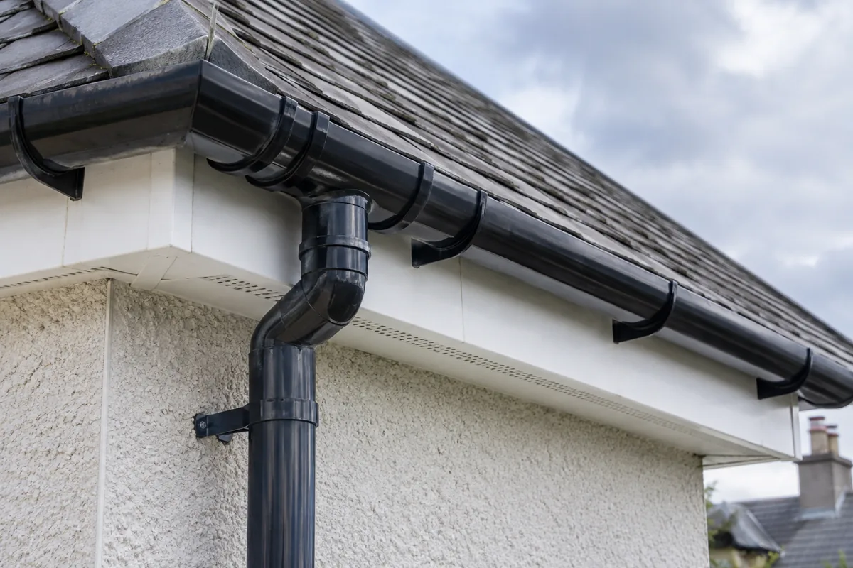 Illustrative gutter outlet and downpipe below white fascia and ventilated soffit.